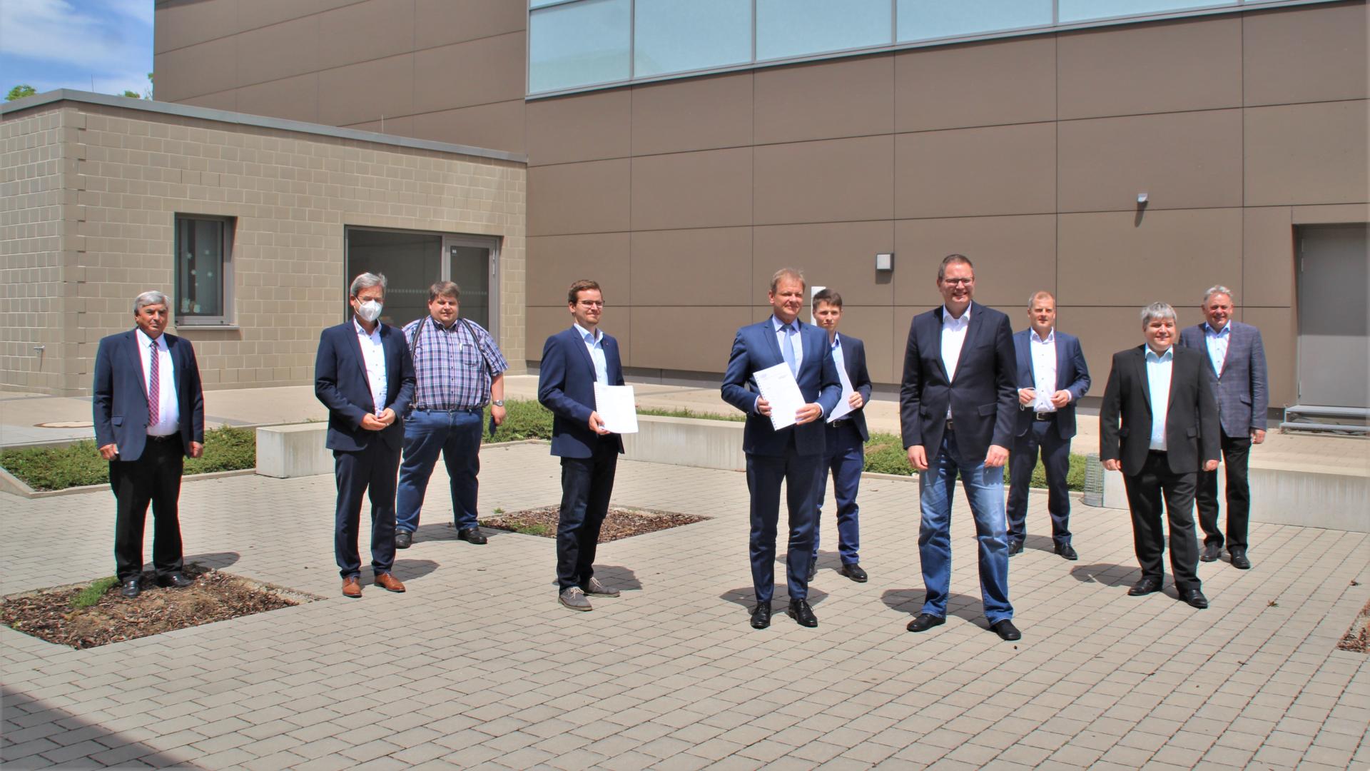 10 men in suits stand in front of a building. Two are holding a document in their hands.