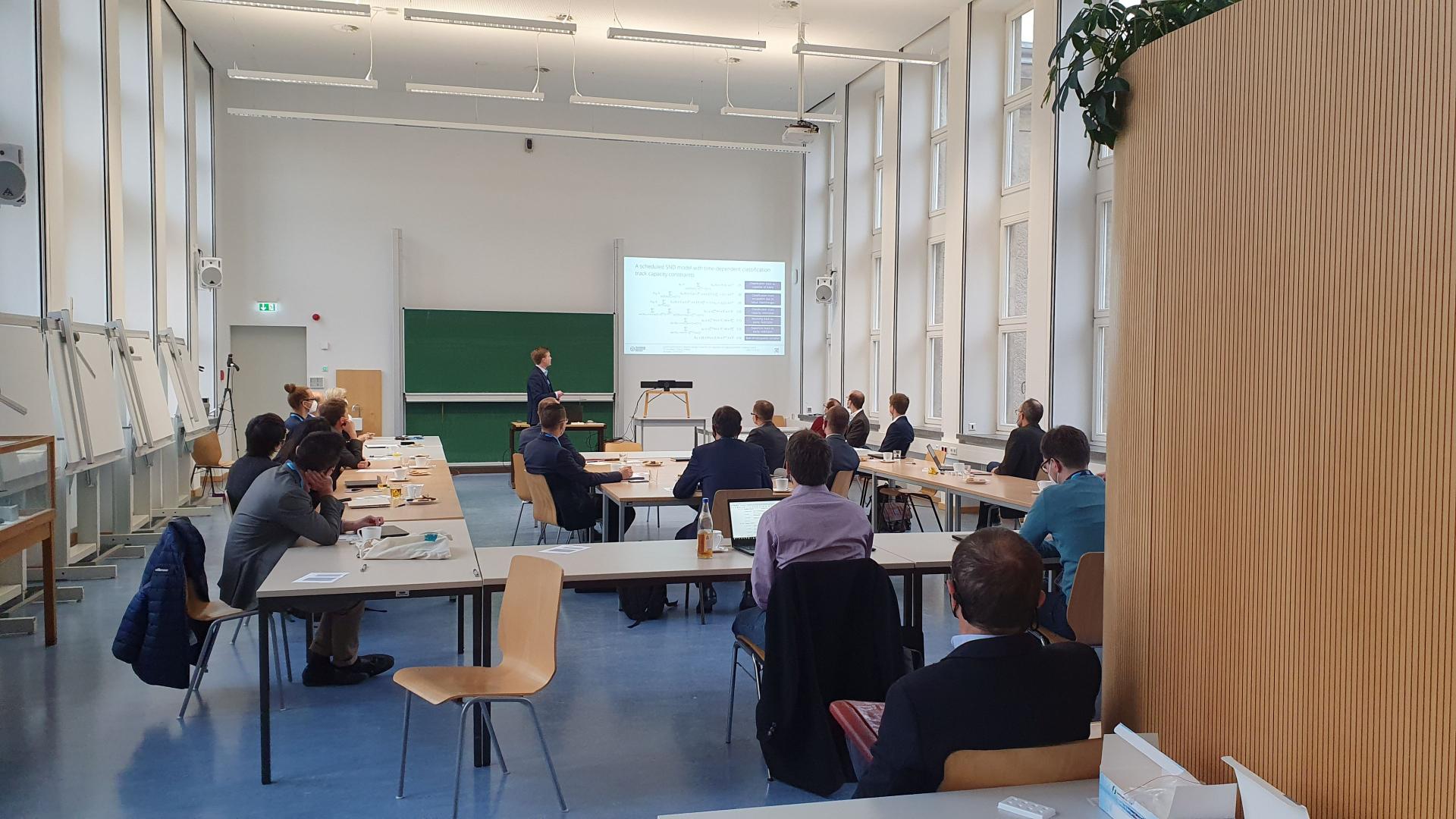 Ein Seminarraum mit Menschen. Einer hält vorn einen Vortrag.