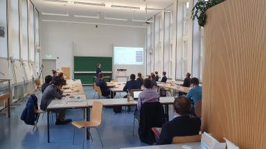 Ein Seminarraum mit Menschen. Einer hält vorn einen Vortrag.