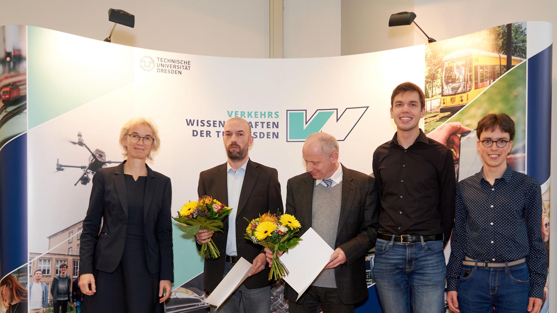 People standing in front of a screen, on the left a woman and then 5 men