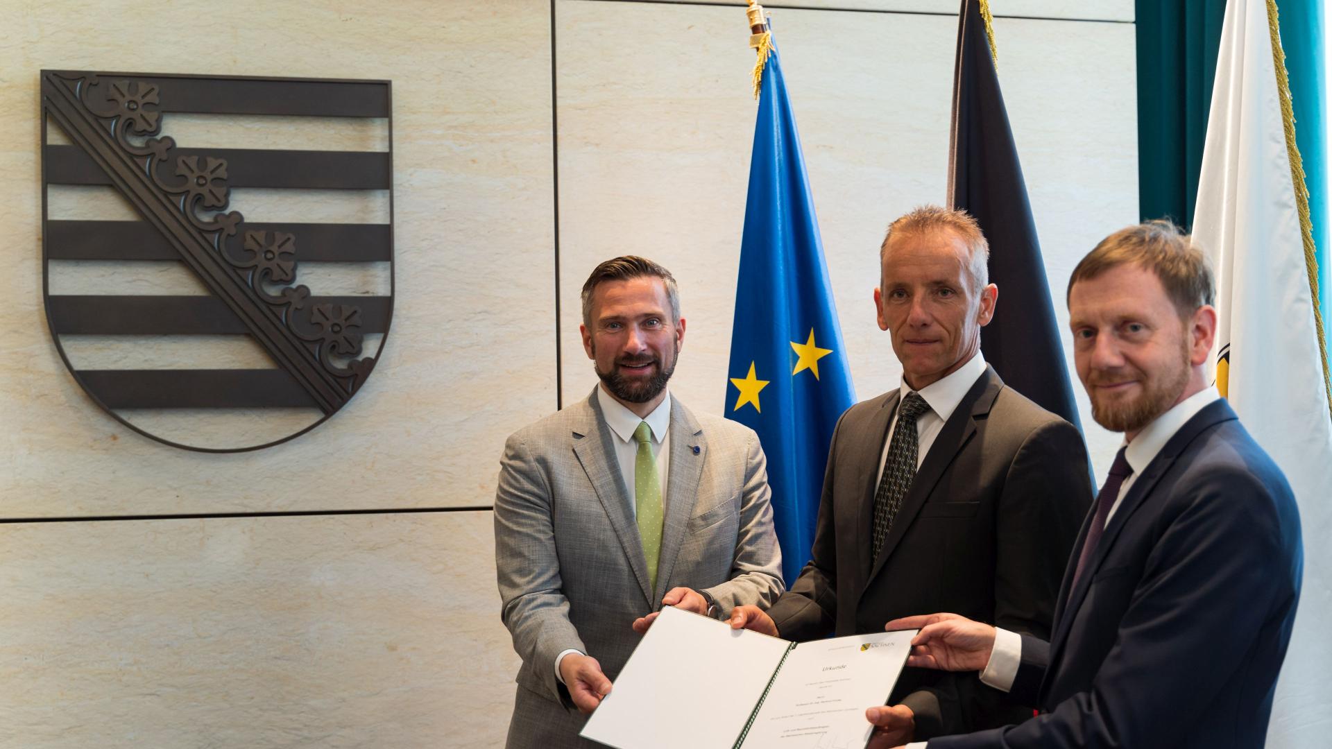 3 men stand side by side holding a certificate together. They are all wearing suits and ties.