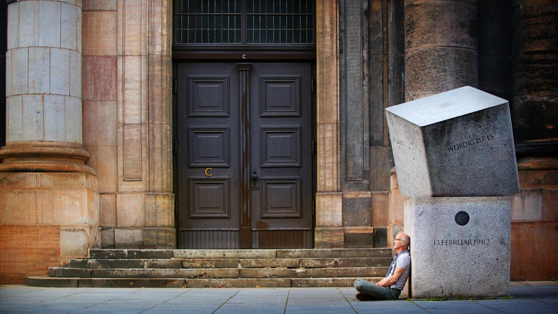 A man sits on the floor, leaning against large granite stones in front of a wooden door.