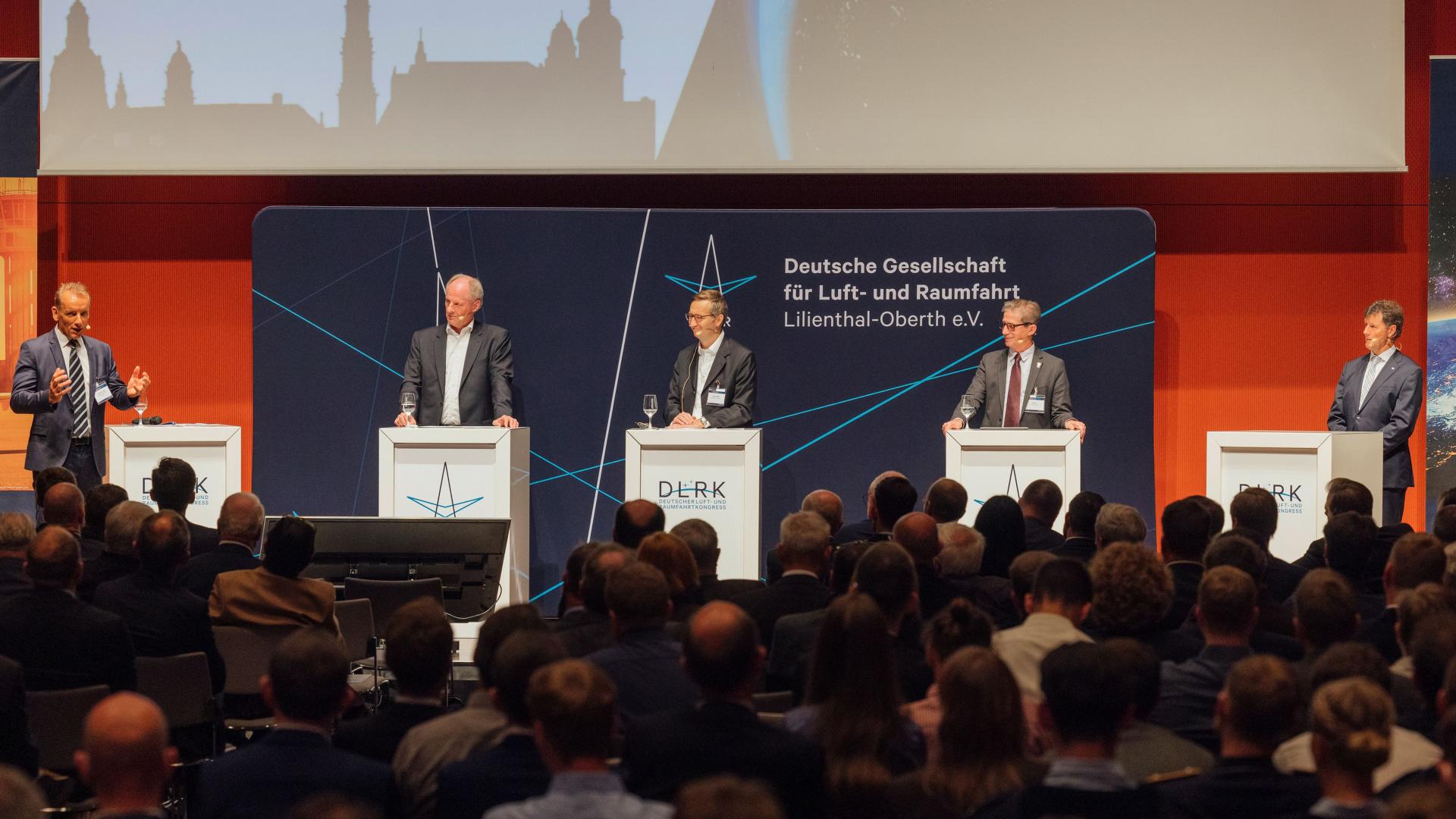 Panel discussion with 5 men in front of a large audience.