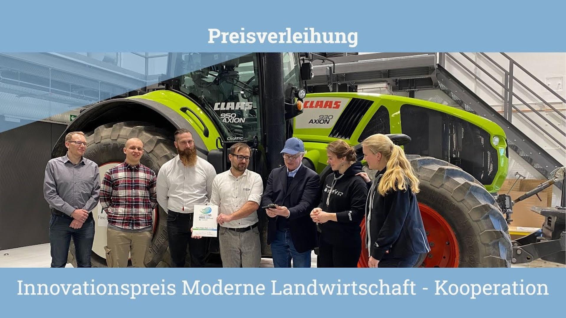 5 men and 2 women stand in front of a tractor. One man is holding a prize, another a certificate.