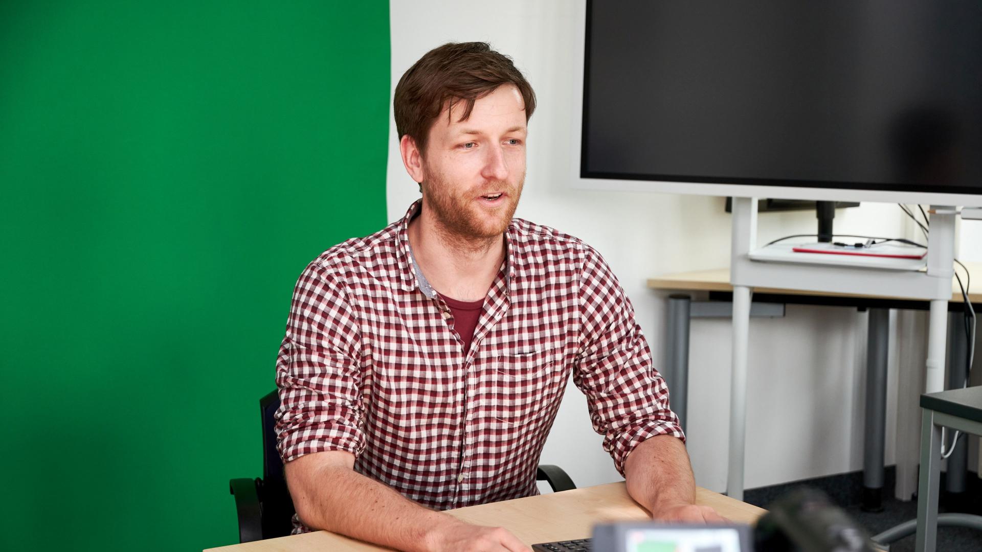 A man sits in front of a camera and a green wall.