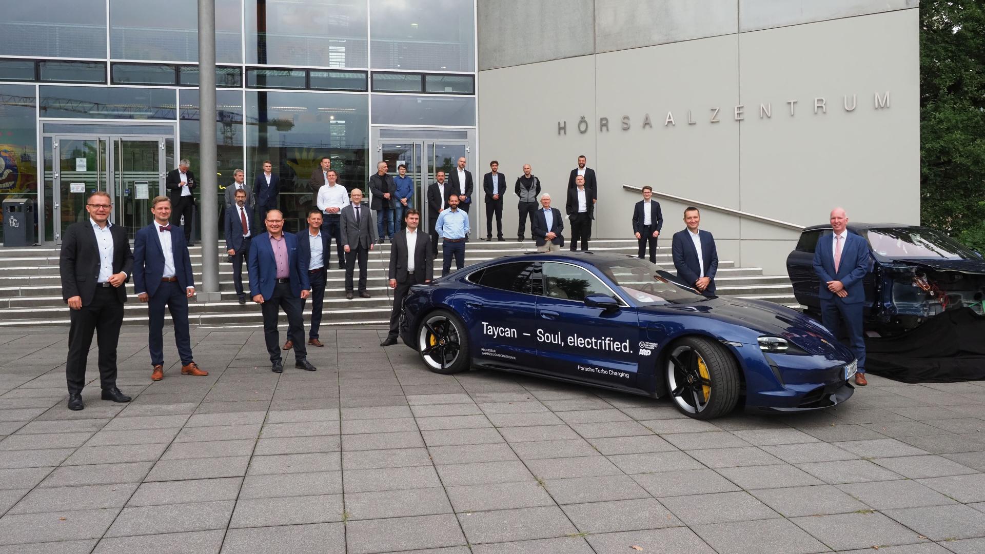 A group of people stand in front of a building, along with a Porsche and a car cockpit.