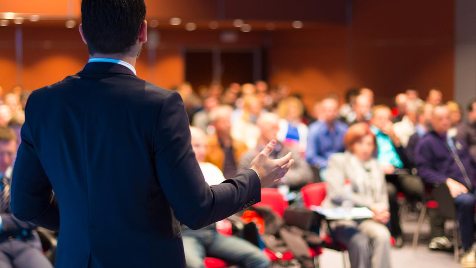 The photo shows a speaker giving a presentation to a large group.