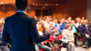 The photo shows a speaker giving a presentation to a large group.
