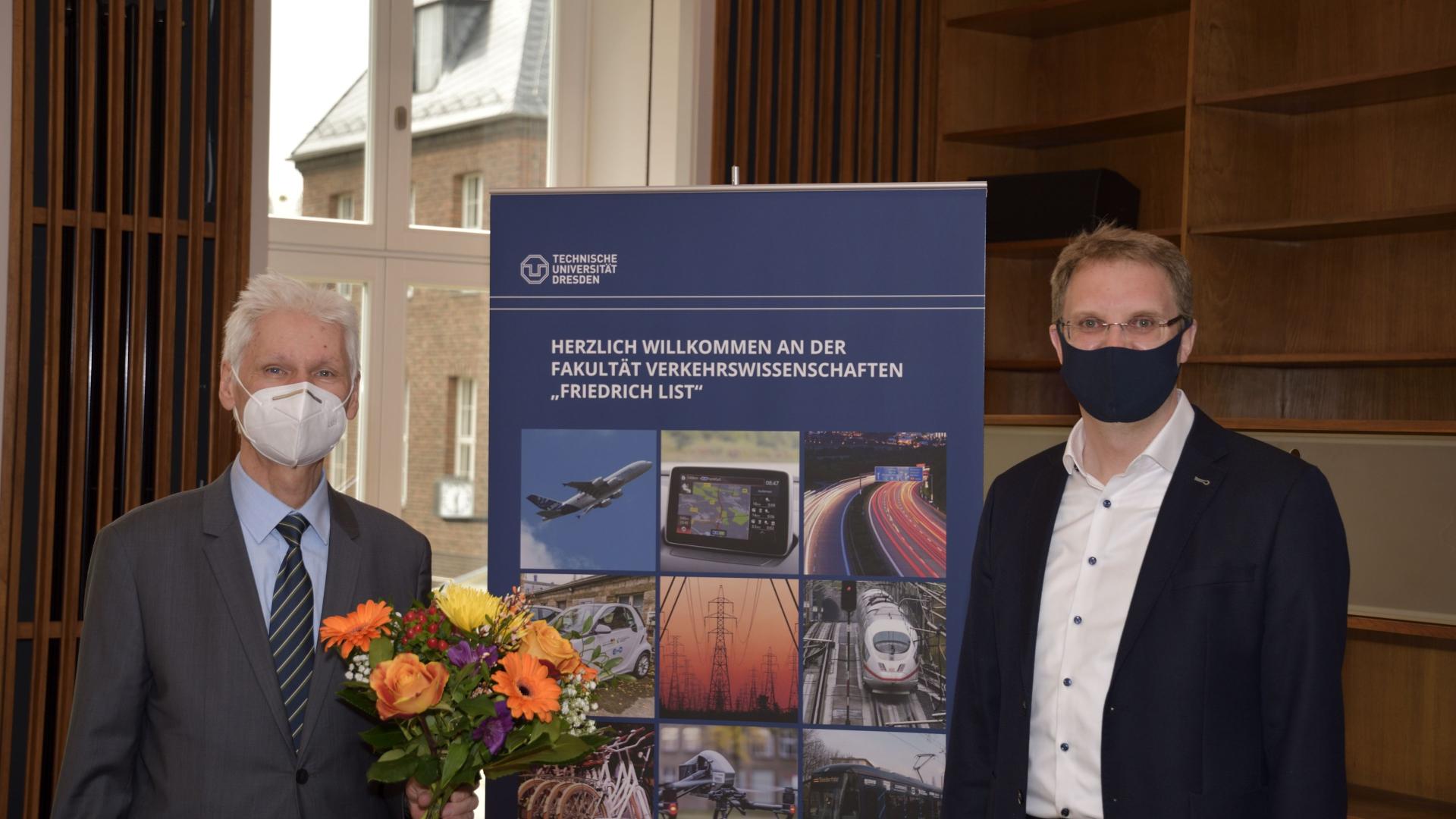Zwei Männder mit Mundschutz nebeneinander, einer hält Blumen, in der Mitte ein Rollup