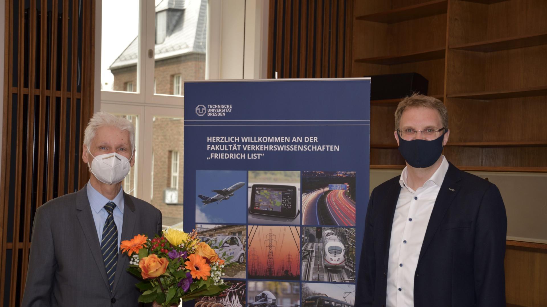 Two men with face masks next to each other, one holding flowers, a rollup in the middle.