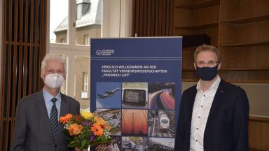 Two men with face masks next to each other, one holding flowers, a rollup in the middle.