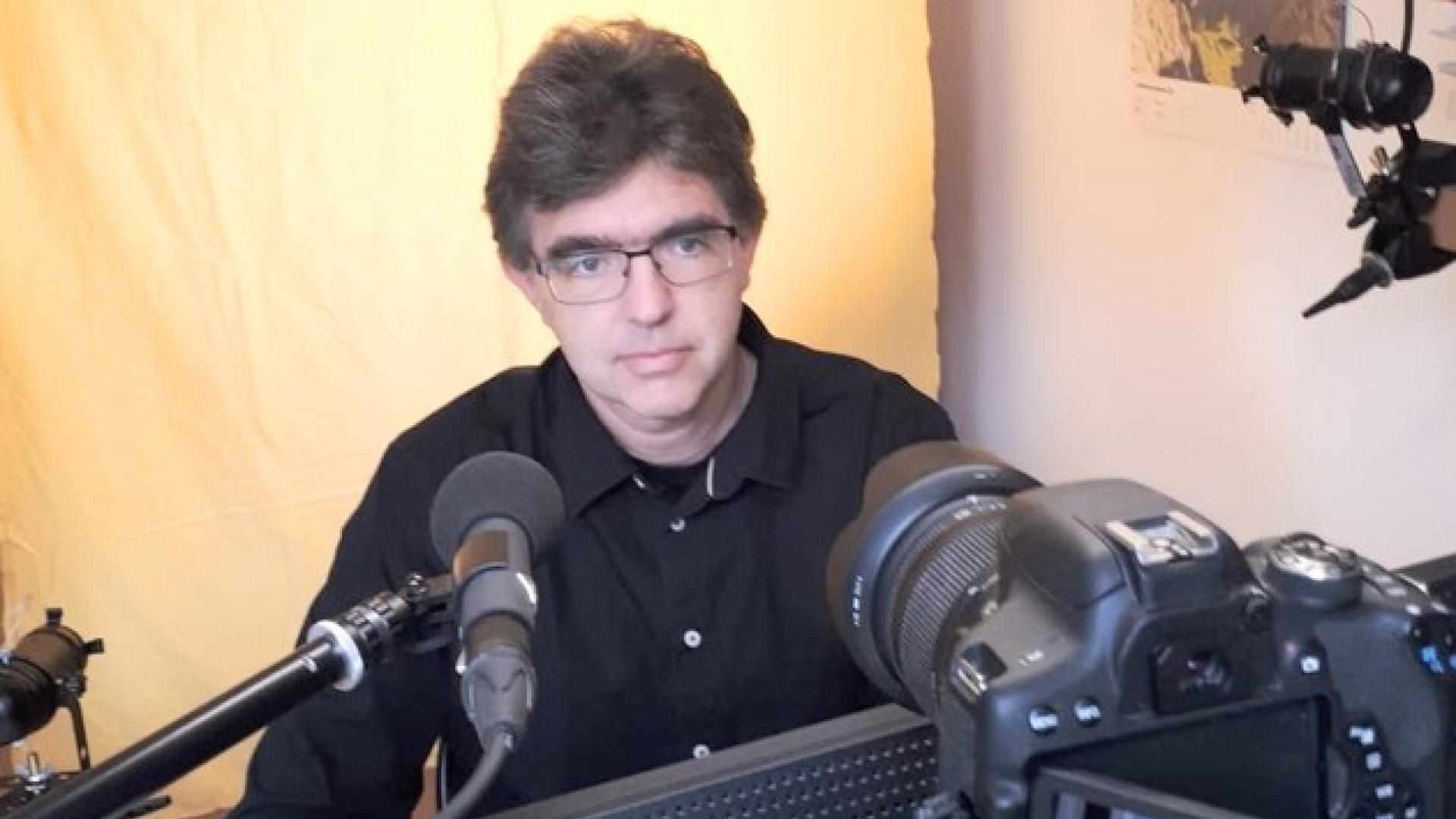 Ein Mann sitzt hinter einem Mikrofon und Laptop. Er trägt eine Brille und ein dunkles Hemd.
