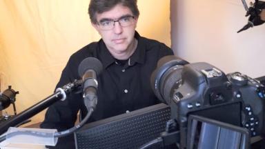 Ein Mann sitzt hinter einem Mikrofon und Laptop. Er trägt eine Brille und ein dunkles Hemd.