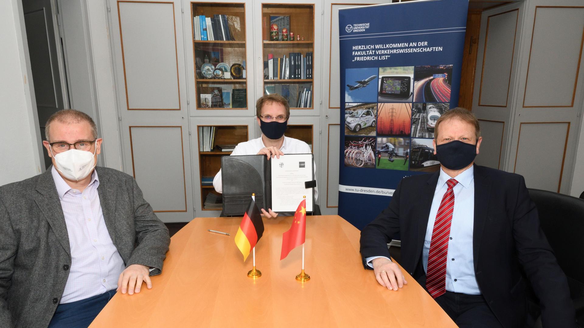 Drei Männer sitzen mit Mundschutz in einem Büro. Der mittlere hält ein Dokument hoch.
