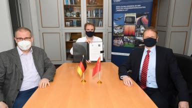 Drei Männer sitzen mit Mundschutz in einem Büro. Der mittlere hält ein Dokument hoch.