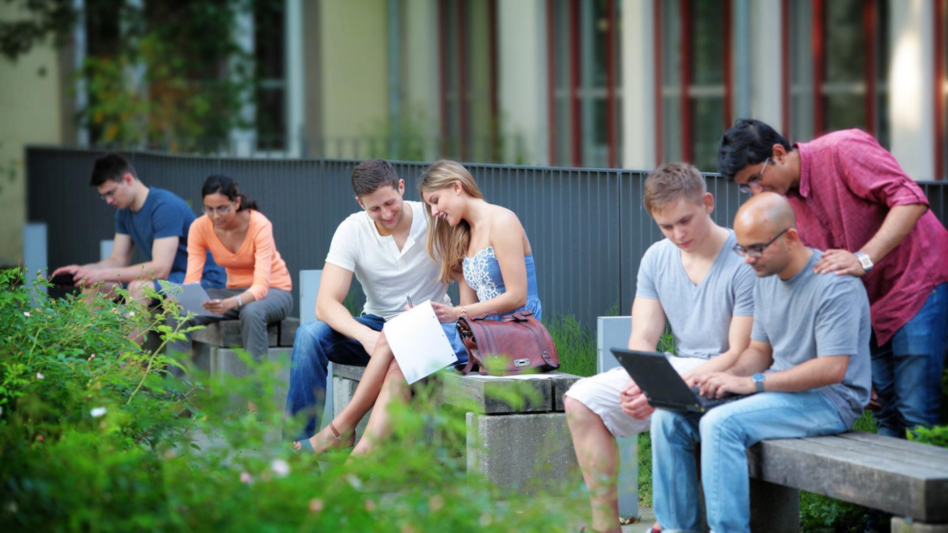 Das Foto zeigt sieben Studierende, die in kleinen Grüppchen auf Bänken im Freien sitzen und lernen.