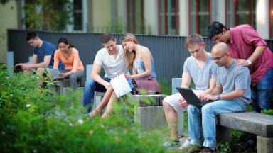 Das Foto zeigt sieben Studierende, die in kleinen Grüppchen auf Bänken im Freien sitzen und lernen.