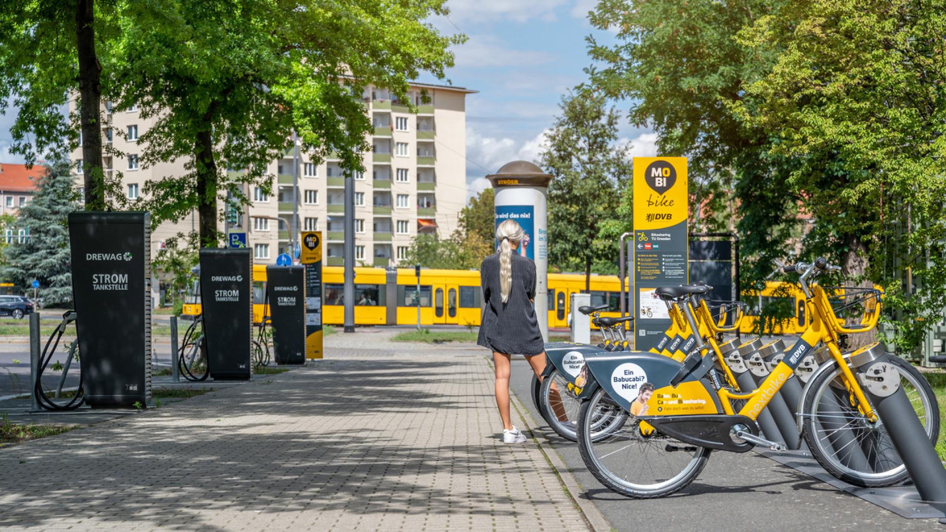 A tram in the background, e-bikes parked on the right, charging stations on the left, a woman