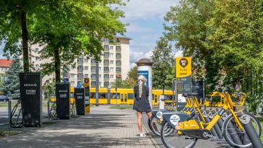 A tram in the background, e-bikes parked on the right, charging stations on the left, a woman