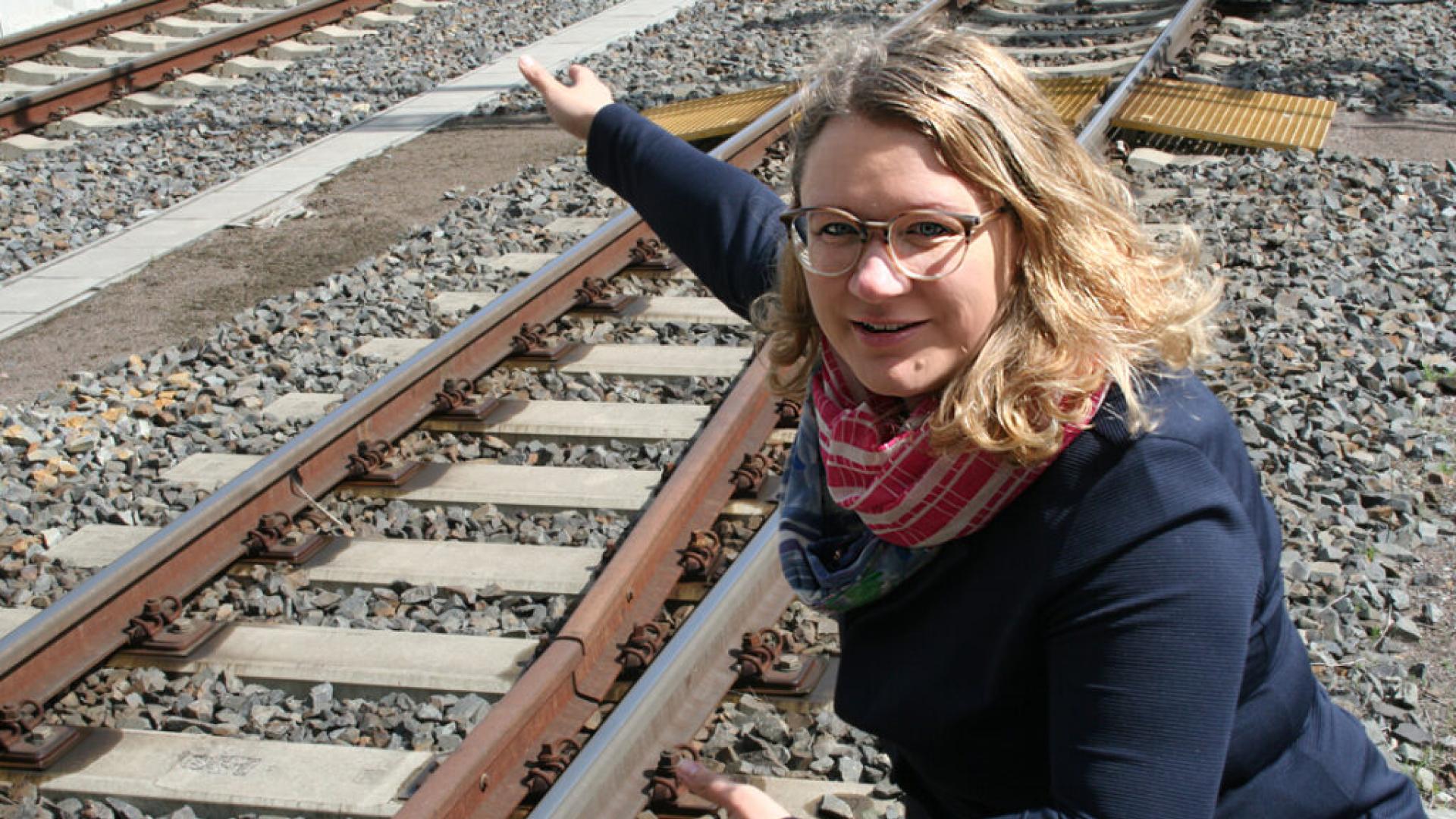 A woman with blond hair and glasses squats by a railway track and points at something.