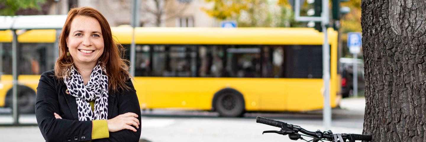 A woman is standing at the side of the road with her arms folded, a bus is driving behind her.