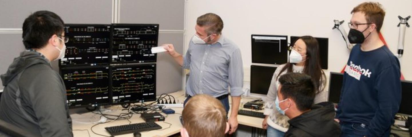  6 People stand and sit in front of a technical device.