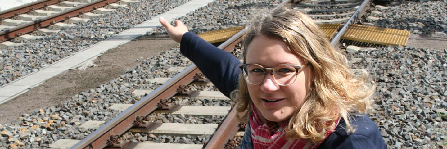 A woman with blond hair and glasses squats by a railway track and points at something.